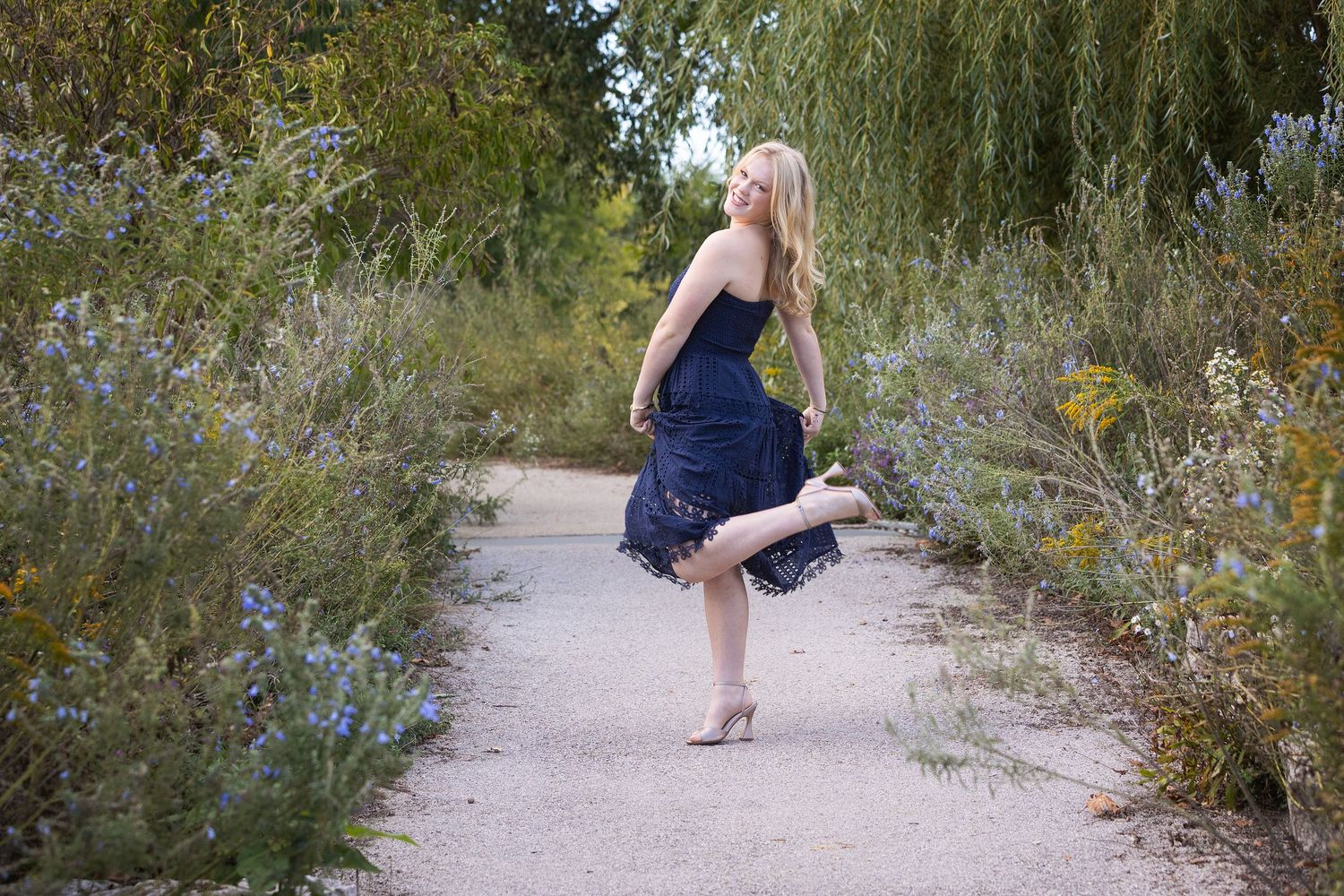 Blonde woman in navy blue dress dancing on a garden path surrounded by lush green trees and wildflowers.