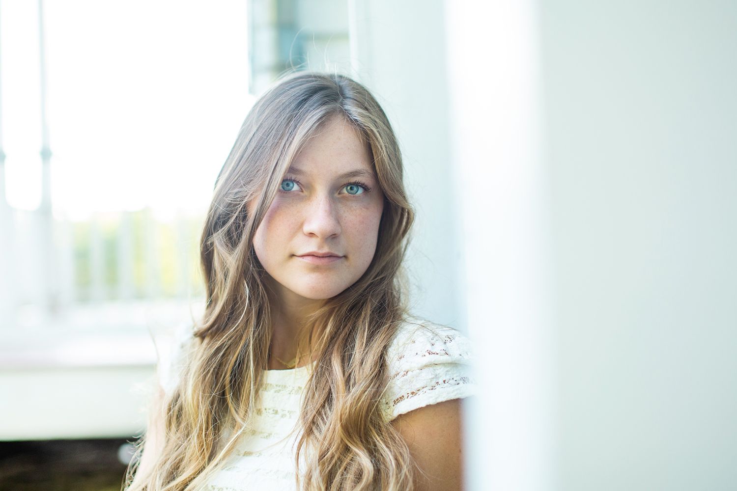 Young woman with long blonde hair and blue eyes wearing a white lace top, gazing thoughtfully near a bright window.