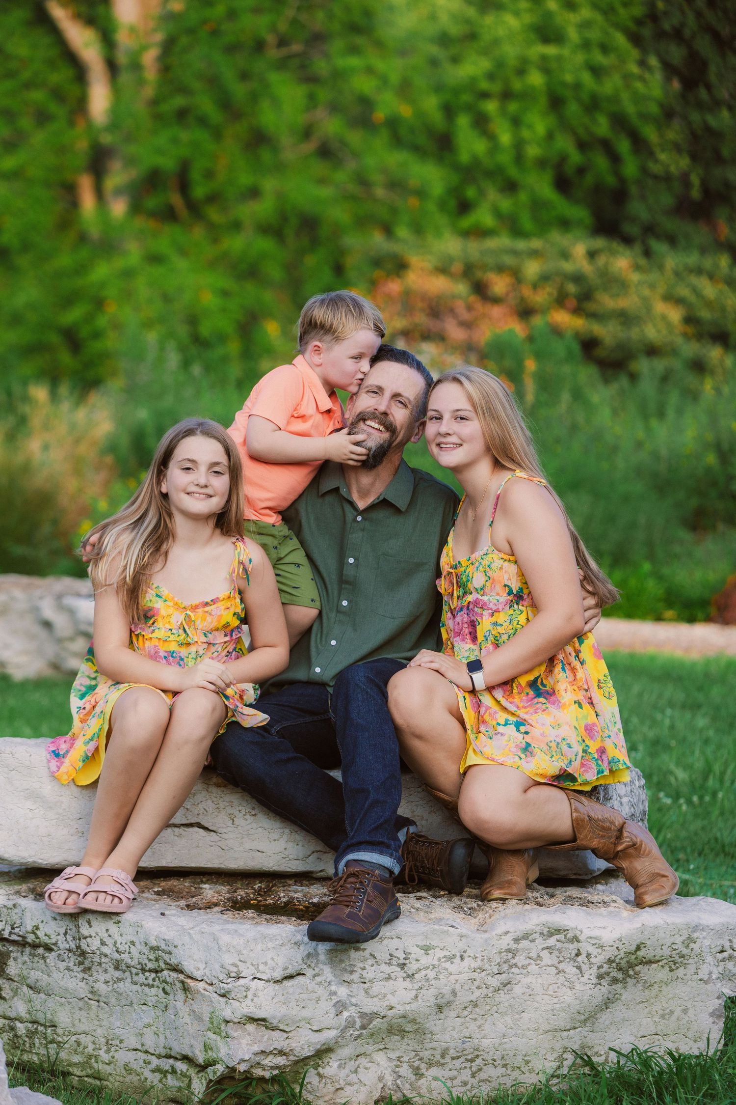 Family of four posing on rocks in a lush green park, wearing colorful summer outfits, smiling and embracing warmly.