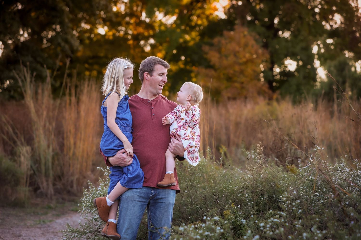A father holds two young daughters in a sunlit field with golden grasses and trees in the background.