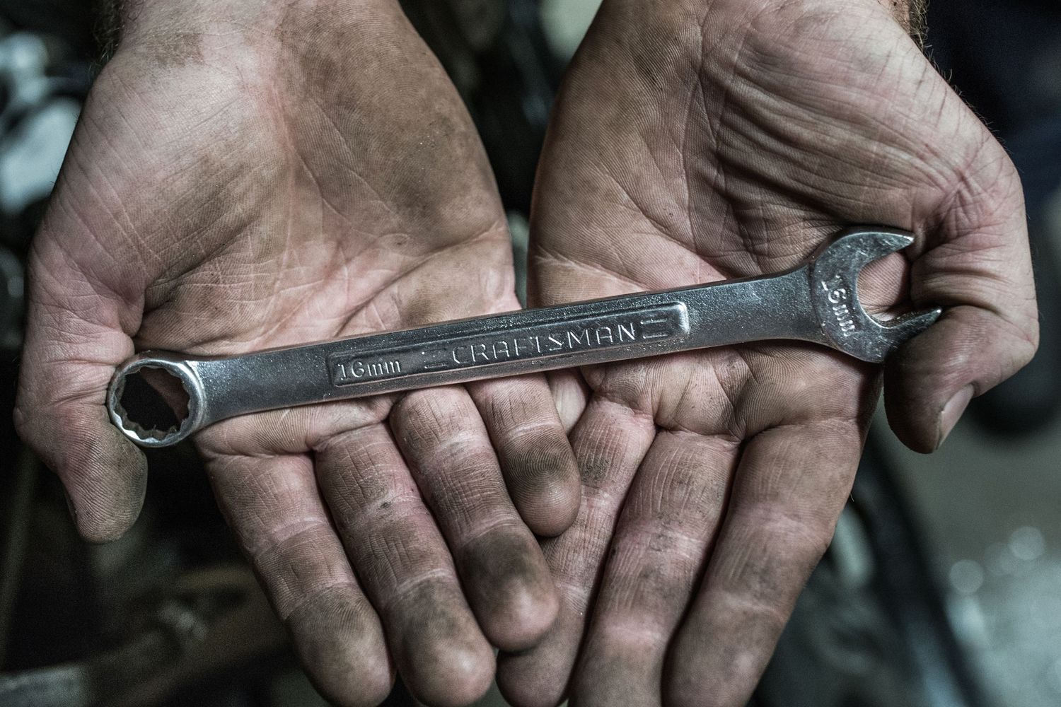 Dirty hands hold a well-worn metal wrench against dark background.