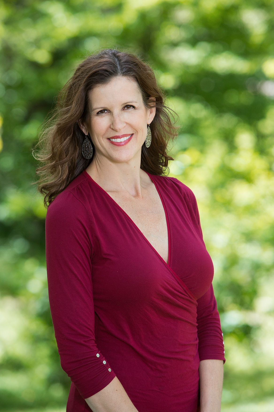Professional portrait of woman in red wrap dress outdoors with natural greenery backdrop.