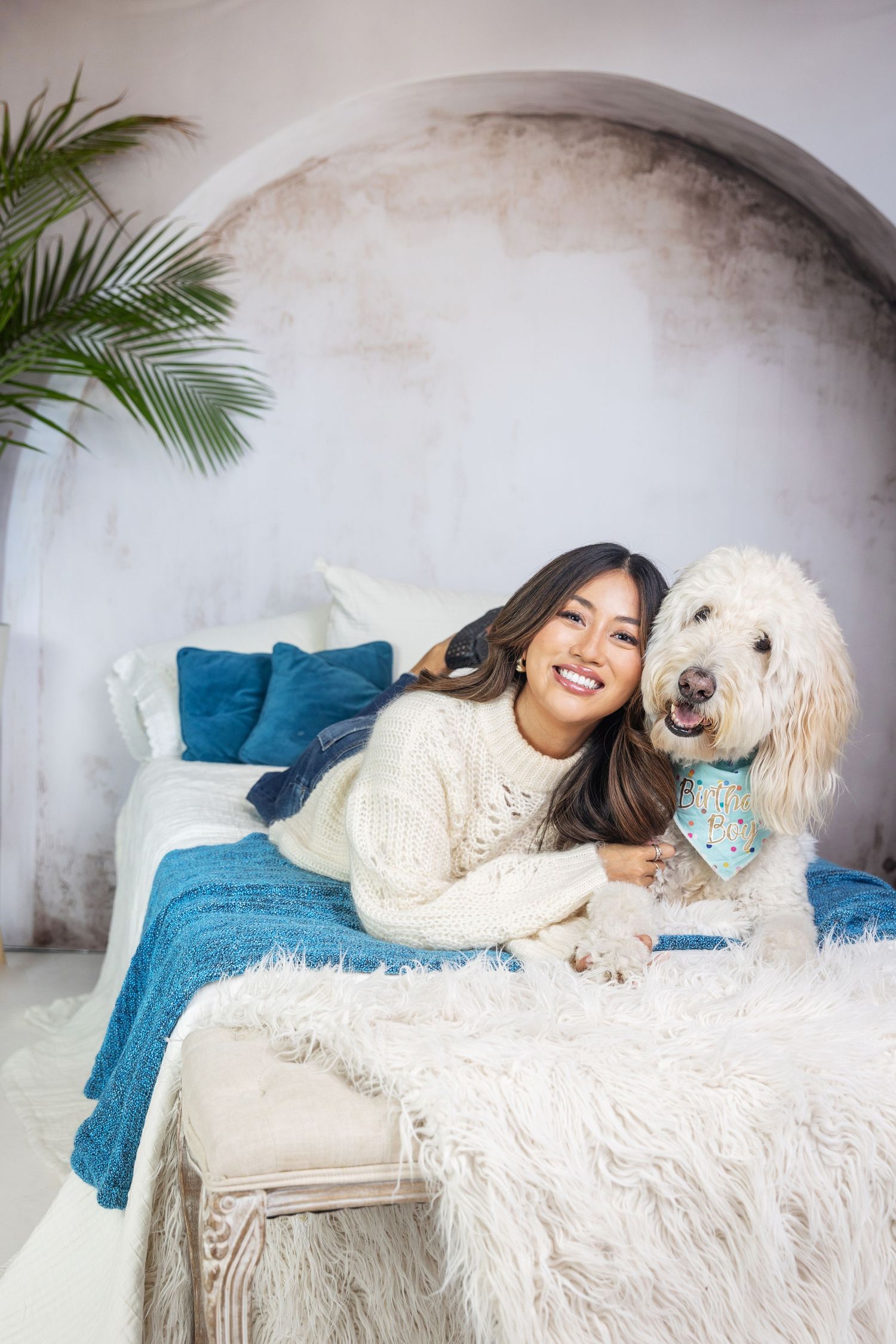 A smiling woman in a white sweater lying on a bed with a fluffy white Goldendoodle wearing a blue bandana.