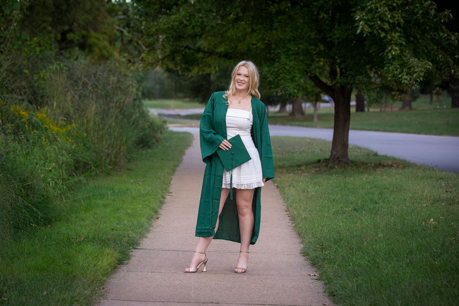 Blonde woman in white dress and green graduation gown holding diploma on tree-lined sidewalk.