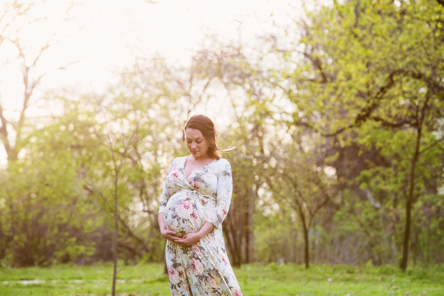 A maternity portrait showing an expecting mother in a floral dress standing in nature at sunset.