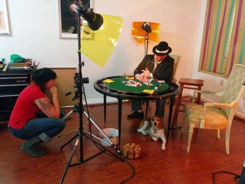 Behind-the-scenes photography setup with dog posing at table while photographer adjusts lighting.