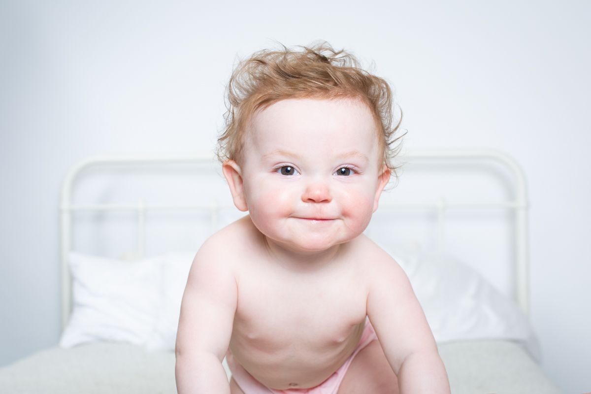 Happy baby sitting on white bedding with rosy cheeks.