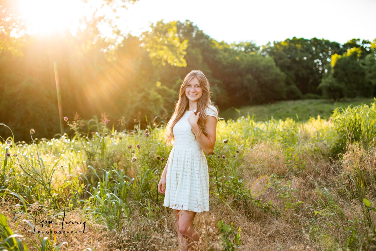 Young woman in white dress standing in golden sunlit meadow with wildflowers and trees during sunset photography session.