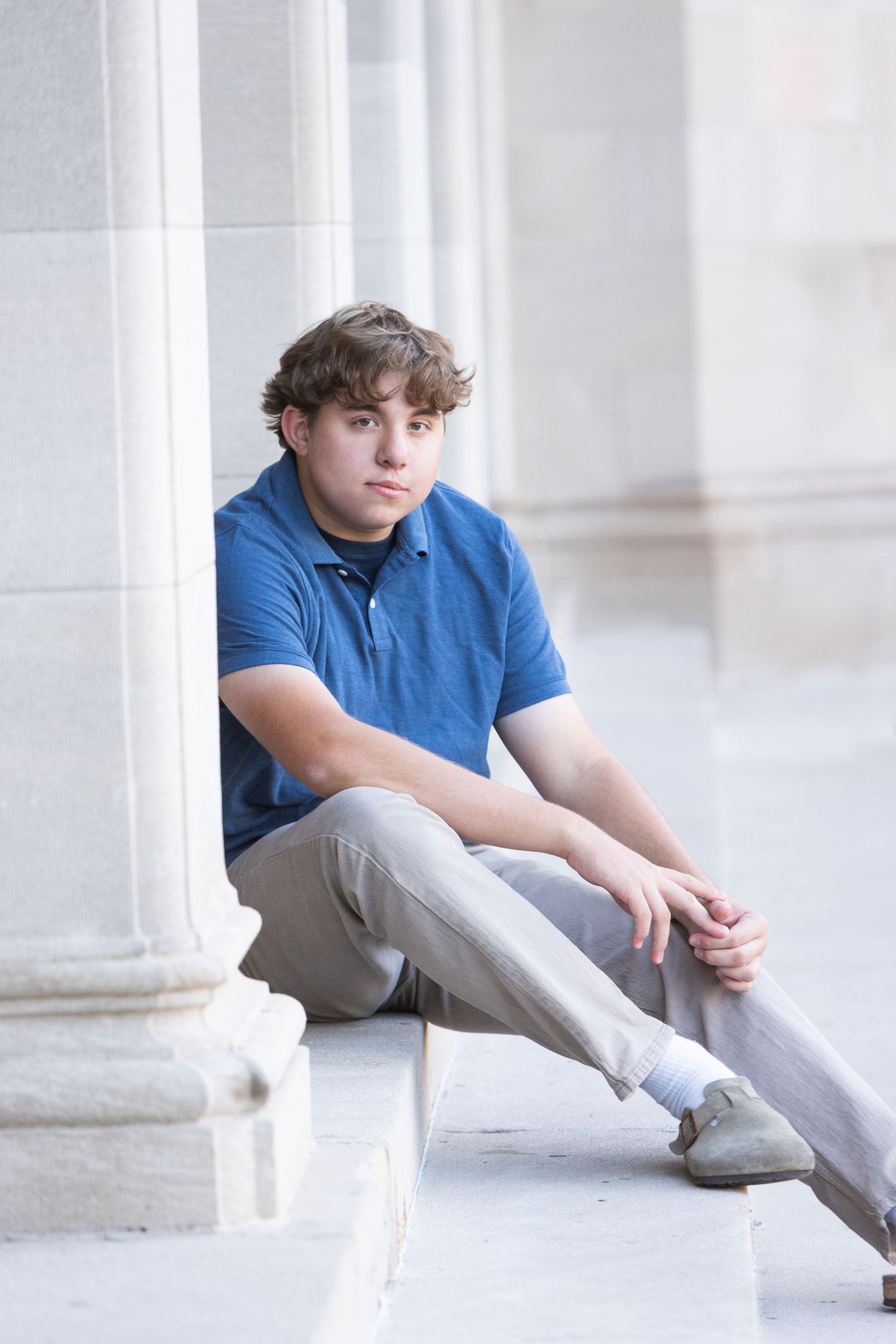 Teen boy in blue polo shirt and khaki pants sitting on stone steps leaning against a column outdoors.