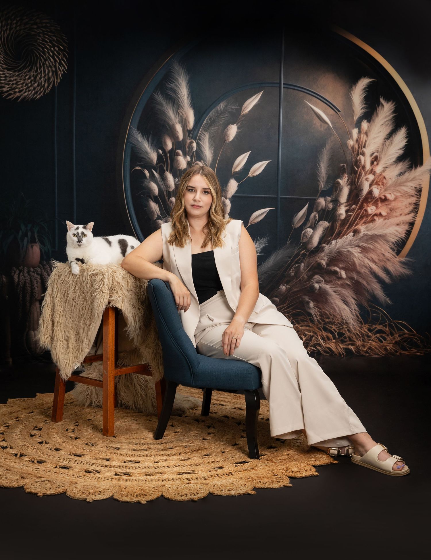 Woman in white outfit sits elegantly beside a fluffy cat on a stool against a dark floral backdrop.
