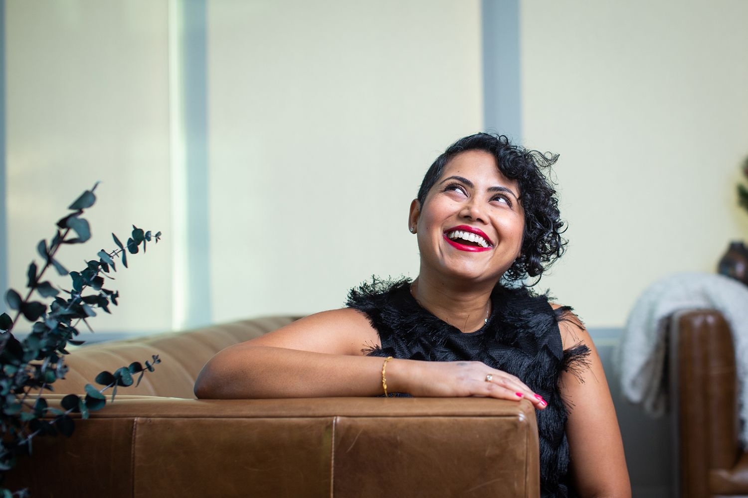 Person smiling and leaning on brown couch with decorative plants in background.
