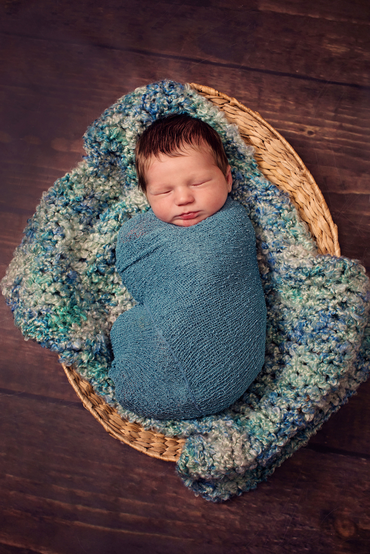 Sleeping newborn wrapped in blue swaddle on textured blanket in wicker basket.