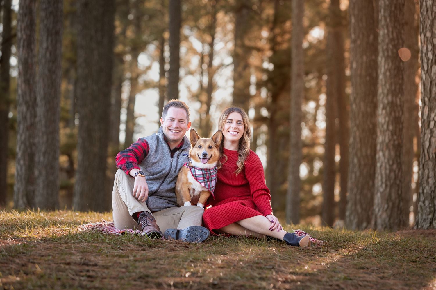 Couple sitting on forest ground with Corgi dog wearing bandana among tall pine trees in warm autumn light.