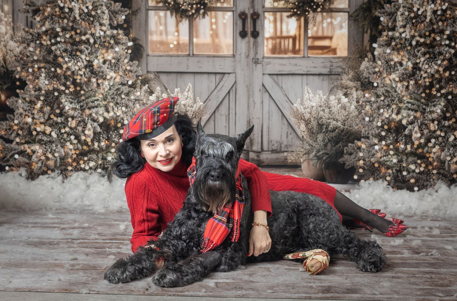 Woman in red outfit poses with large black Schnauzer dog in festive Christmas photo with decorated trees and barn doors.
