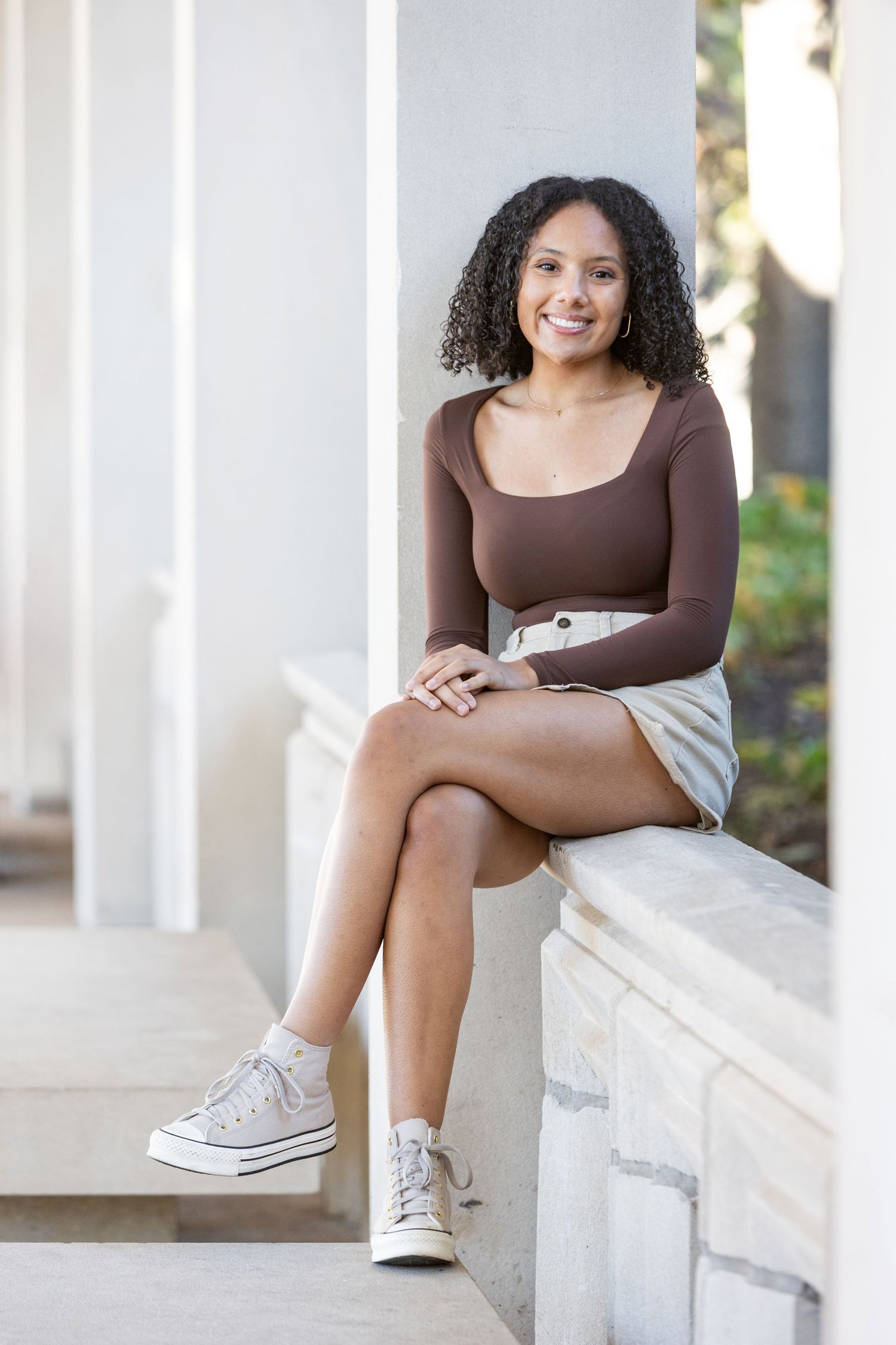 Young woman in brown top and mini skirt sits smiling on stone ledge, wearing white Converse sneakers.
