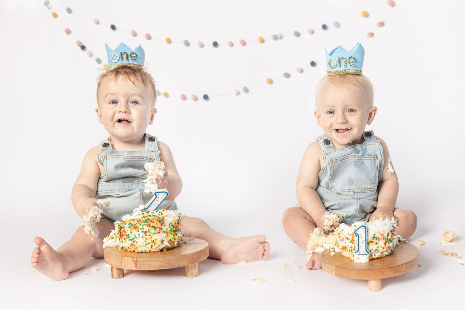 Twin babies in denim rompers celebrate their first birthday with smash cakes, wearing blue crown hats on white background.