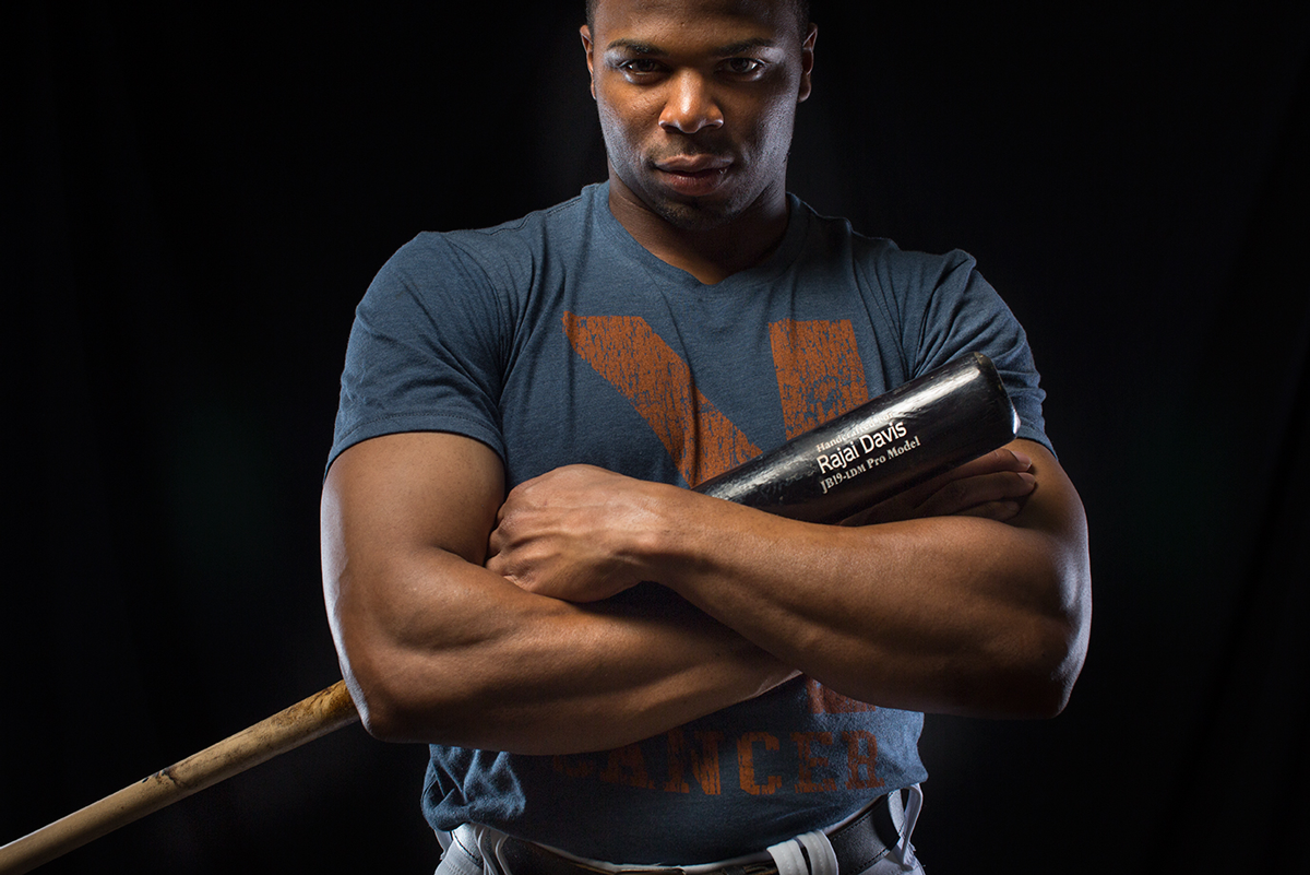 Athletic figure in dark t-shirt poses with crossed arms holding baseball bat against black background.
