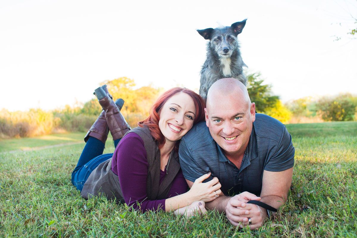 A playful outdoor portrait with a small dog perched atop a couple lying in the grass.