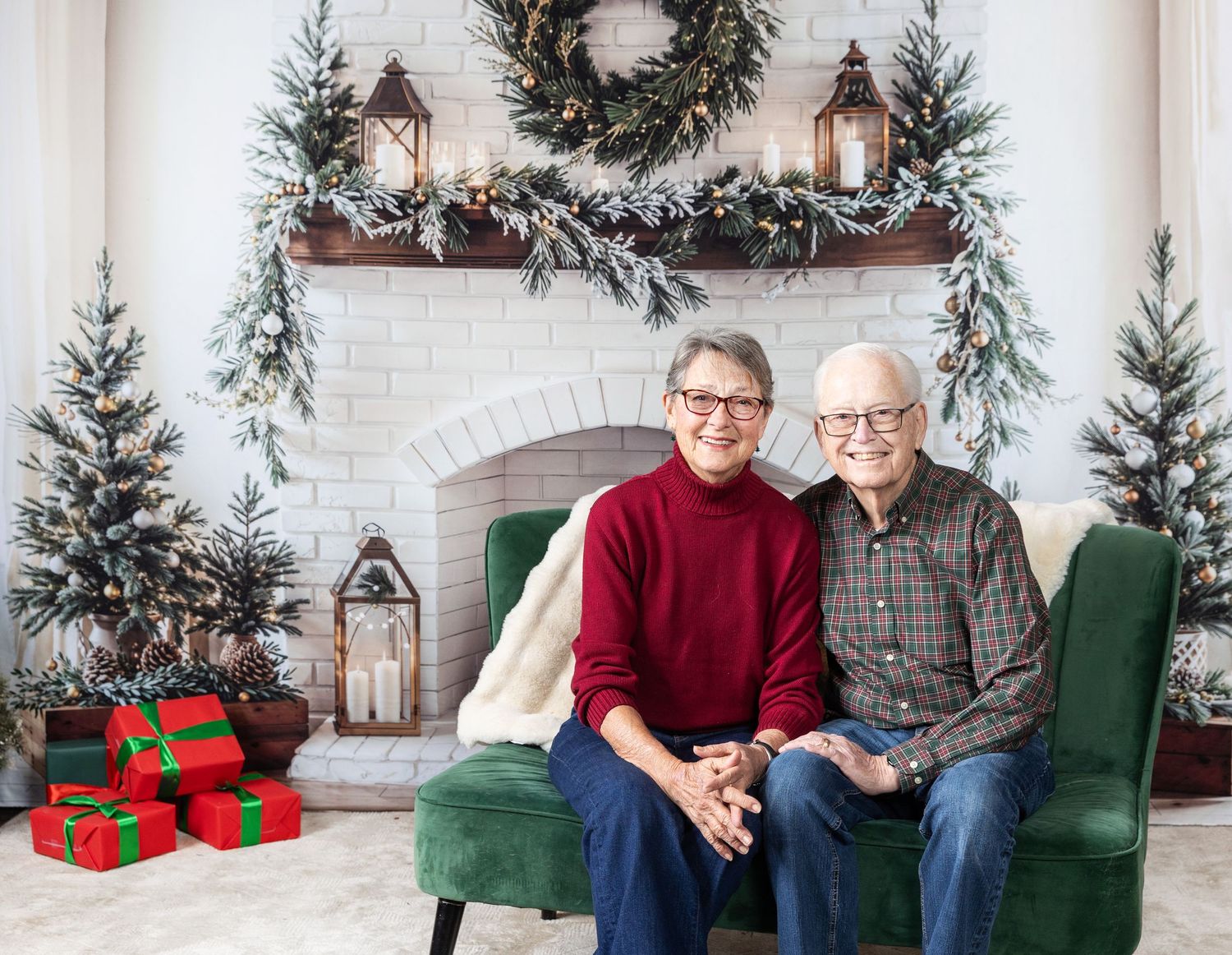 Elderly couple smiling together on green velvet chair by decorated Christmas fireplace with wrapped gifts and holiday trees.