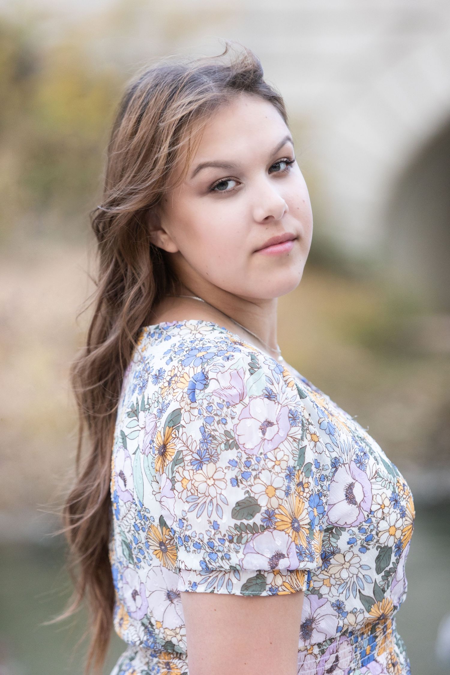 Young woman in floral dress with long brown hair posing outdoors near a river with a soft bokeh background.