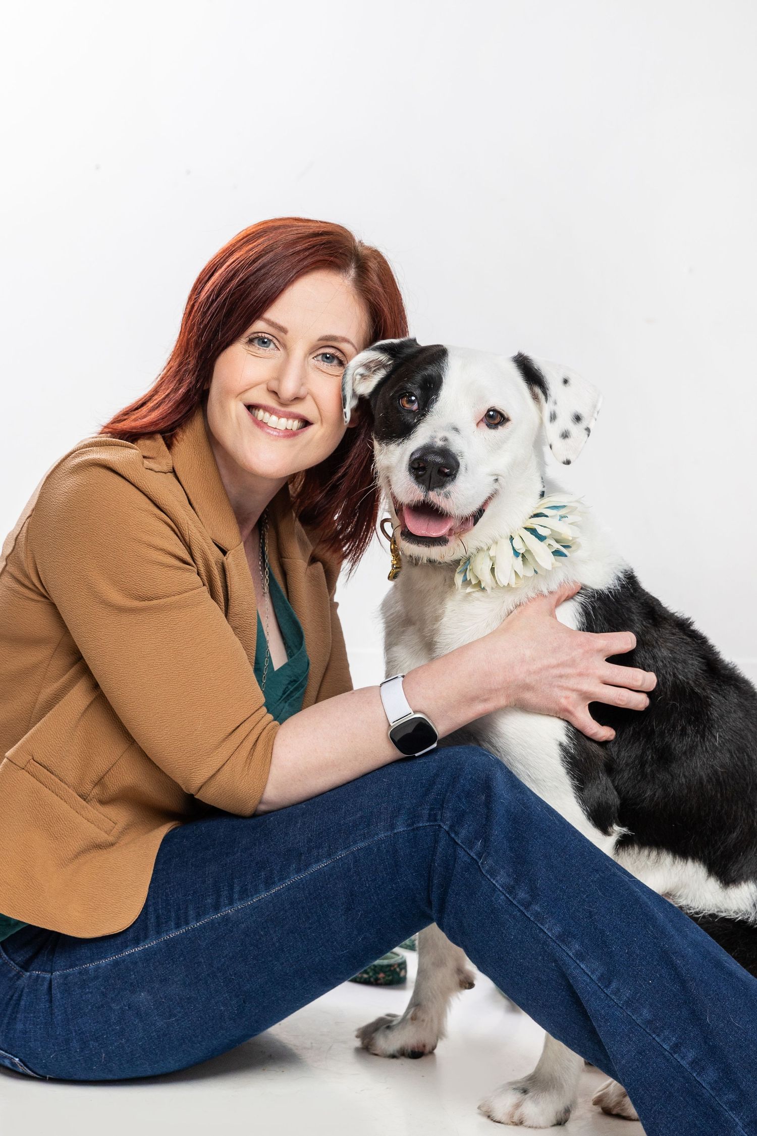 Smiling redhead woman in brown blazer and jeans sits with black and white dog against white background.
