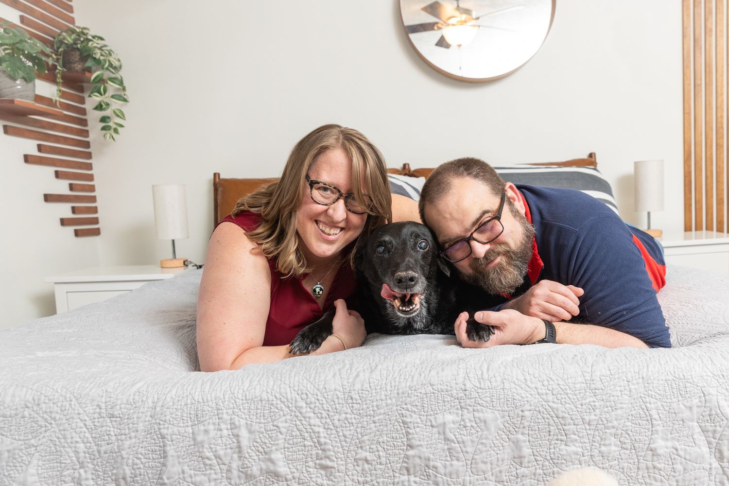 Smiling couple with glasses lying on a bed with their happy black dog in a cozy bedroom.