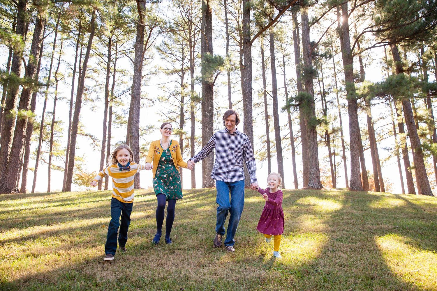 Family of four holding hands and walking through a sunny pine forest on green grass.