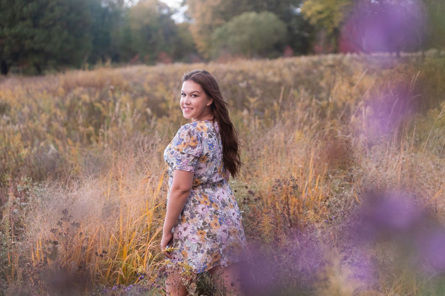 A young woman in a floral dress smiles over her shoulder in a golden meadow with purple wildflowers.