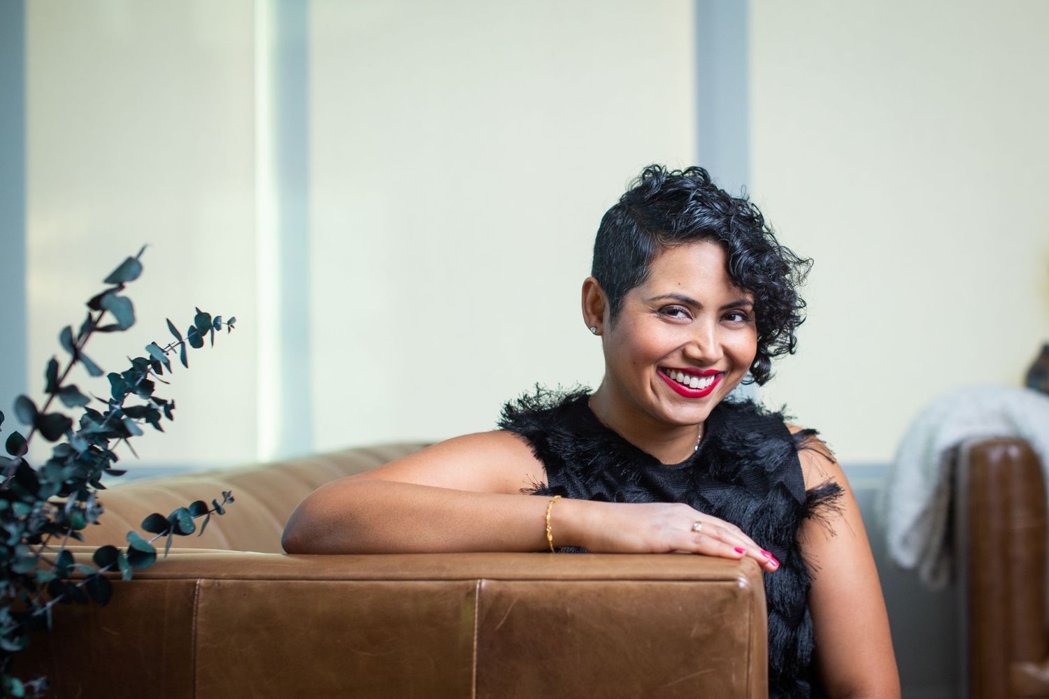 A person in a black textured dress relaxes on a brown leather couch with plants in the background.