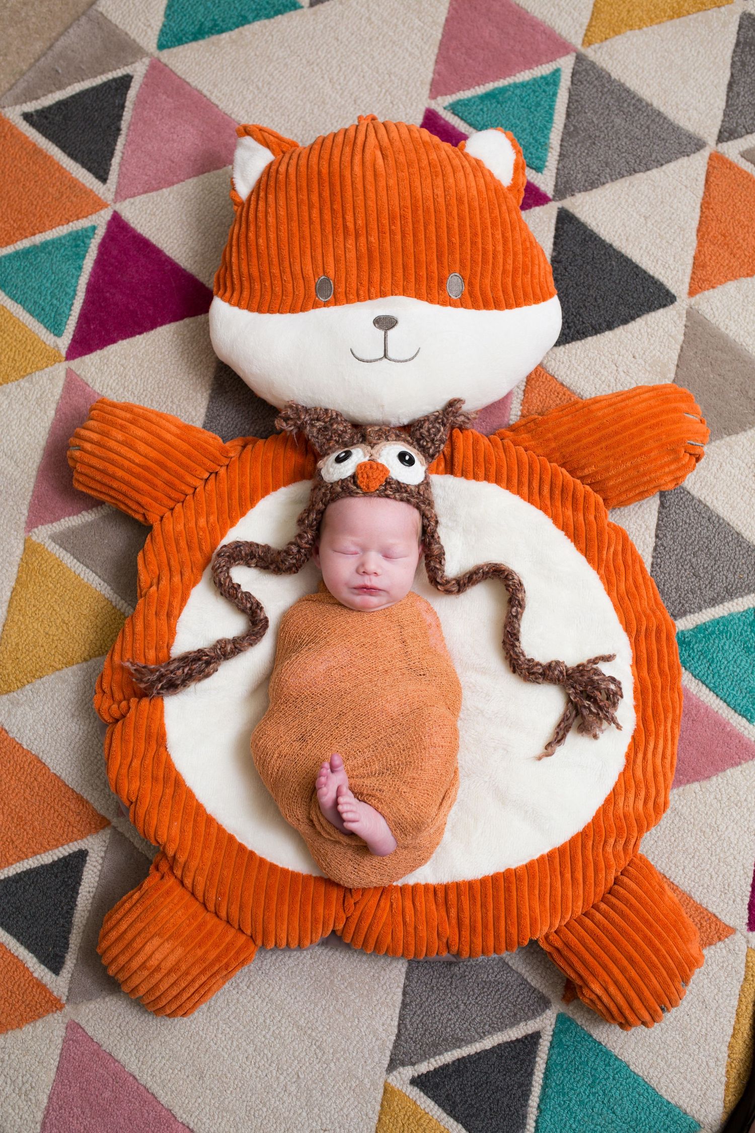 A small baby in brown swaddle lying on an orange and white fox-shaped plush mat with geometric print carpet beneath.