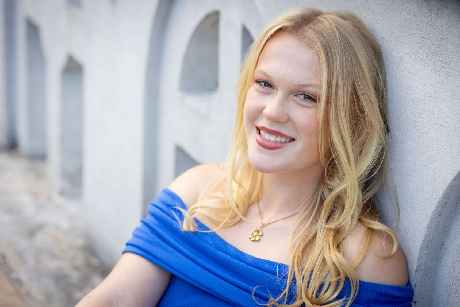 Smiling young woman with blonde hair wearing a blue off-shoulder top and gold anchor necklace, leaning against a wall.