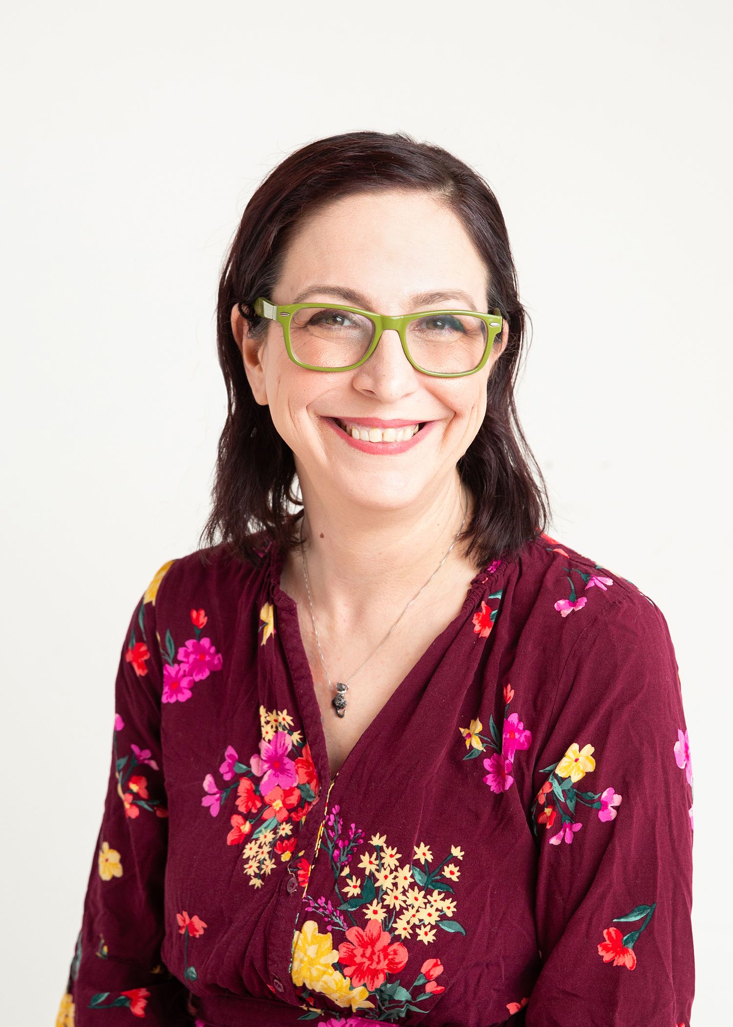 Woman wearing stylish glasses and floral burgundy top in professional headshot against white background.