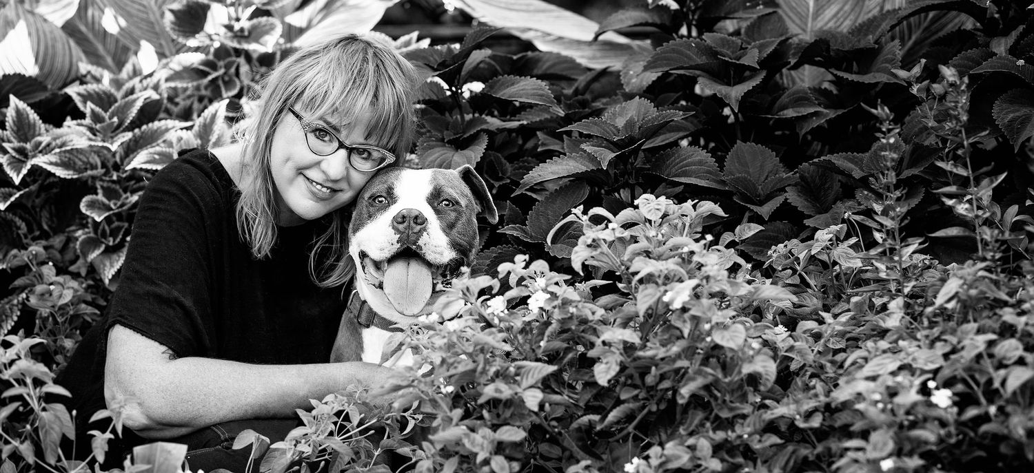 Black and white portrait of a pit bull and companion among leafy foliage.