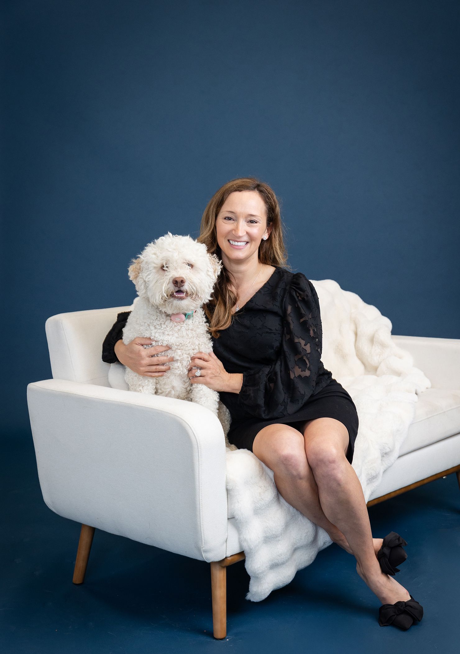 A smiling woman in a black dress sits on a white sofa with a fluffy white dog against a blue background.