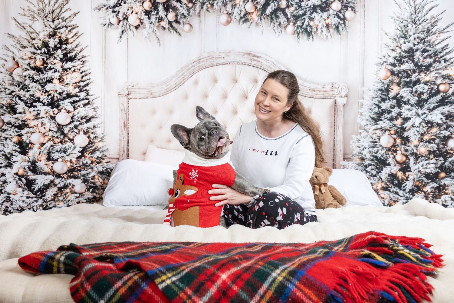 Woman in pajamas smiling with French Bulldog holding Christmas gift on bed with festive trees.