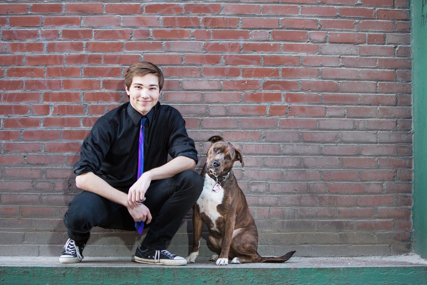 Young man in black shirt and blue tie squatting beside a brown dog in front of a red brick wall.
