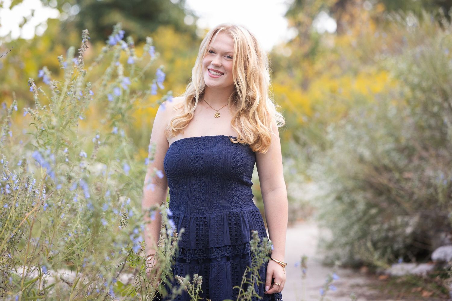 Smiling blonde woman in navy strapless dress standing among wildflowers in a sunlit outdoor garden setting.