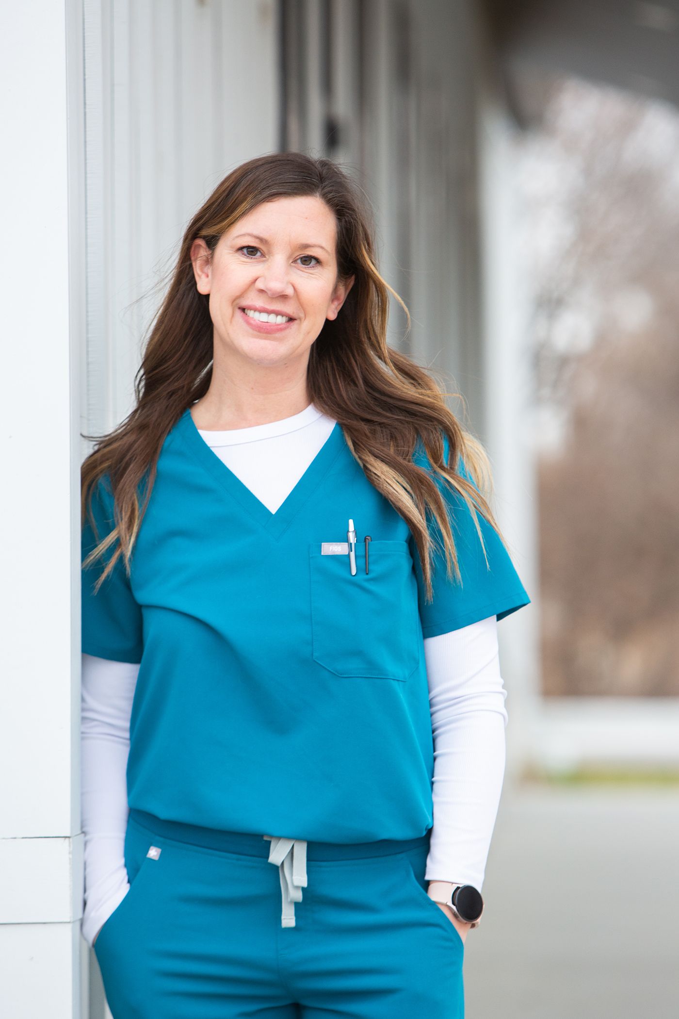 Healthcare worker in teal scrubs smiles brightly while standing outside medical facility.