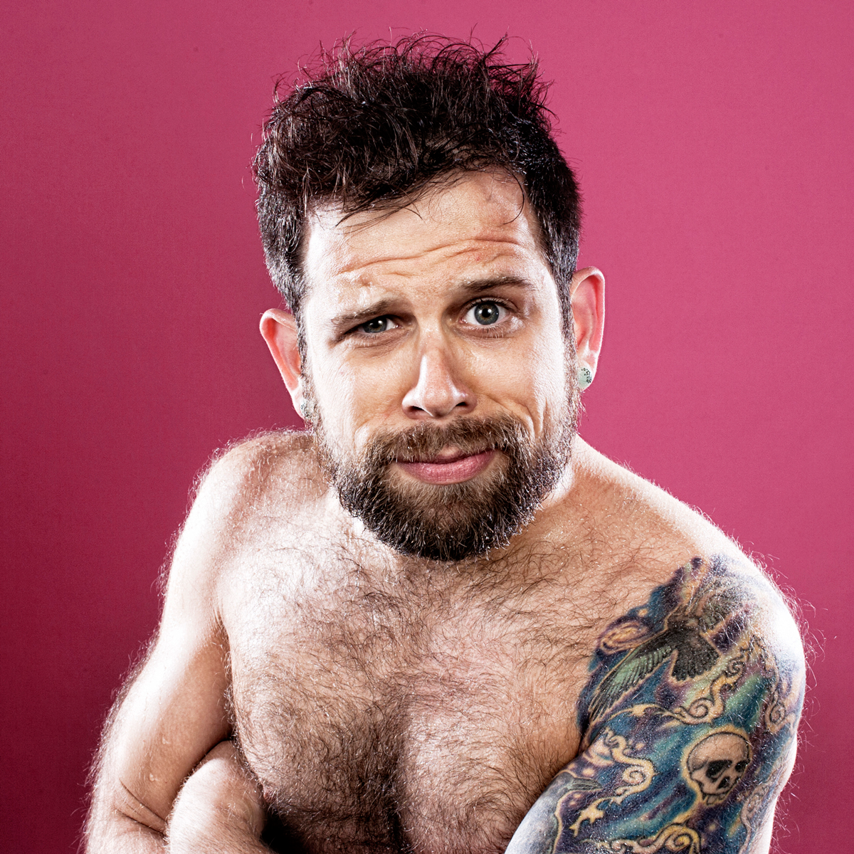 Portrait against pink background showing shirtless person with beard and colorful tattoo sleeve.