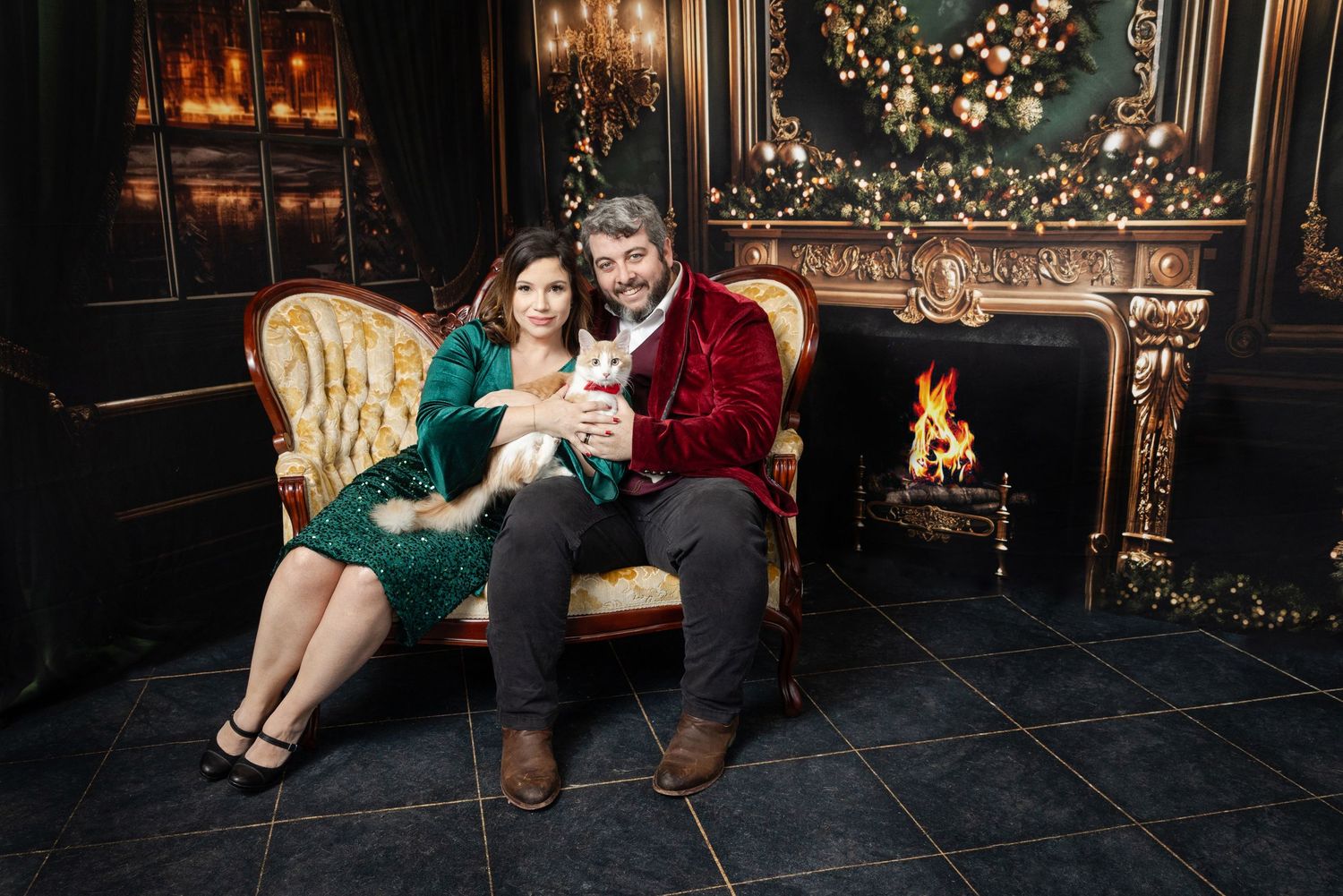 Couple sitting on a gold sofa by a fireplace with a small dog, decorated for Christmas.