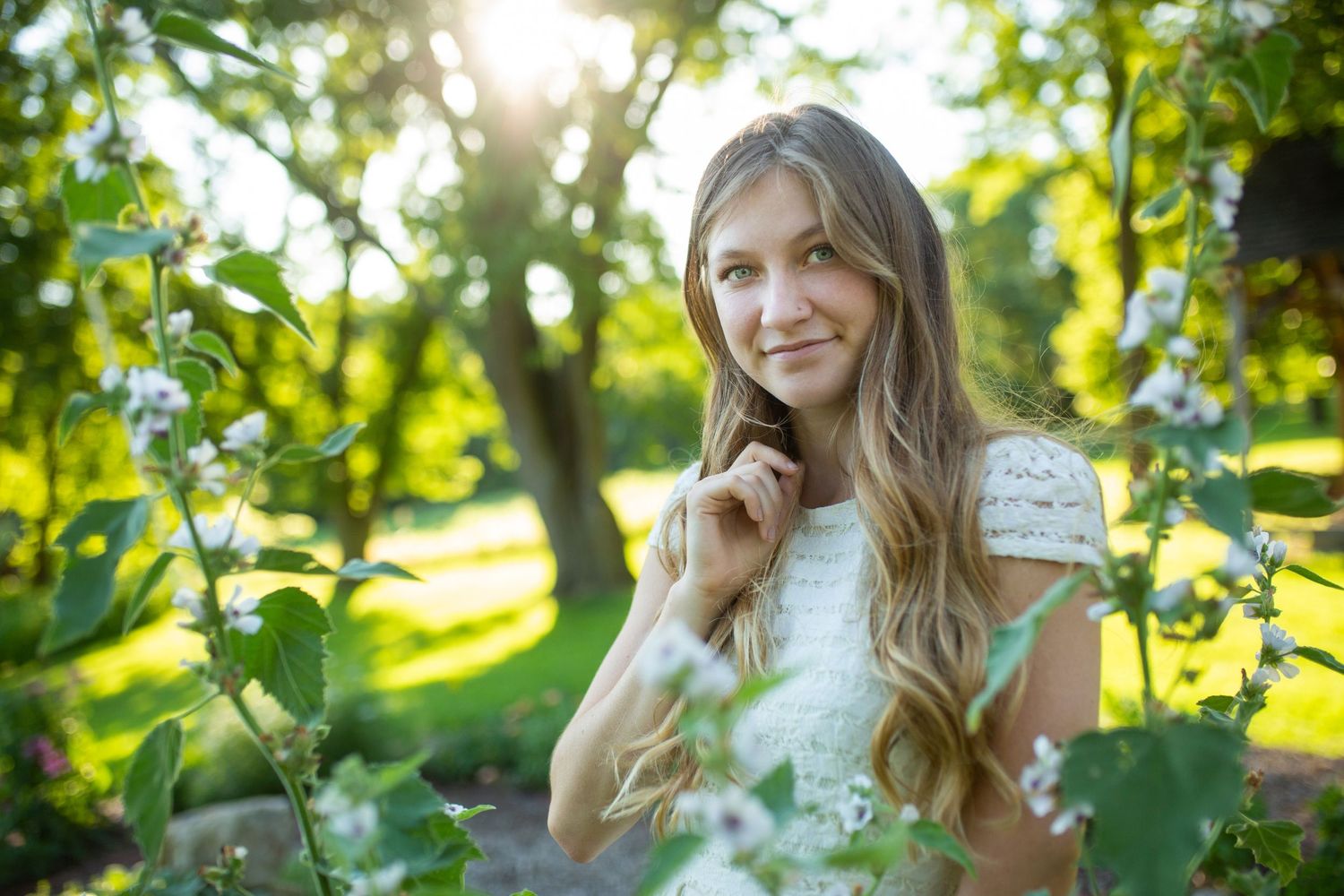 Young woman with long blonde hair smiling in a sunny garden surrounded by blooming white flowers and green trees.