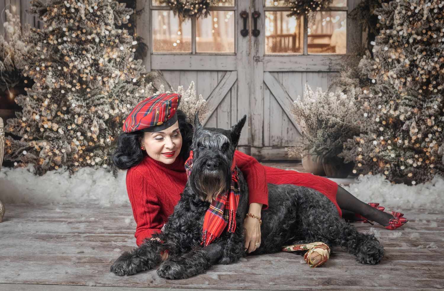 Woman in red dress and tartan hat poses with black Schnauzer dog near festive Christmas barn backdrop.