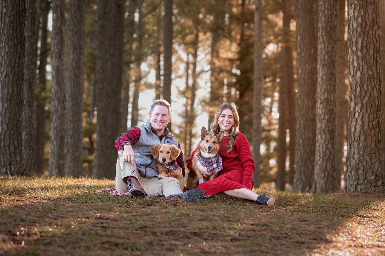 Couple sitting outdoors in a pine forest with two dogs, wearing red and plaid outfits in a fall family portrait.