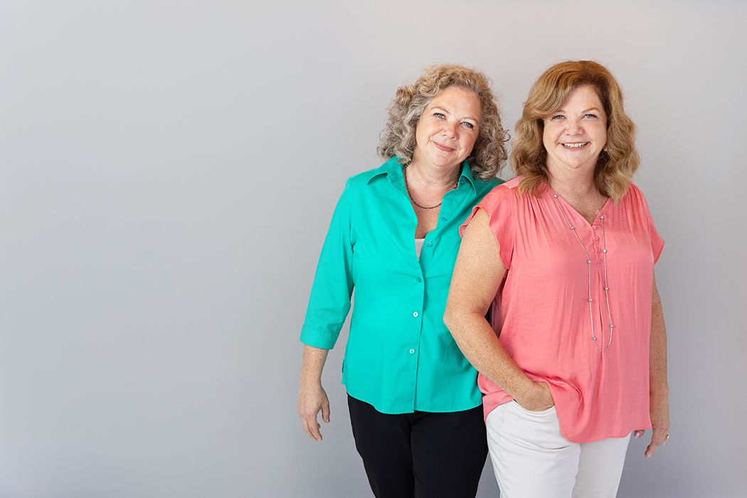 Two women in turquoise and coral tops smile together against light grey wall.