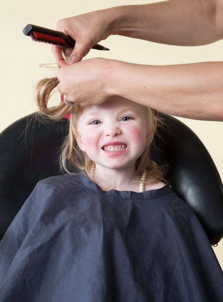 Young child getting a haircut while sitting in a salon chair and smiling.