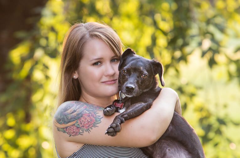 A person with tattoos hugging their black dog outdoors with yellow foliage in background.