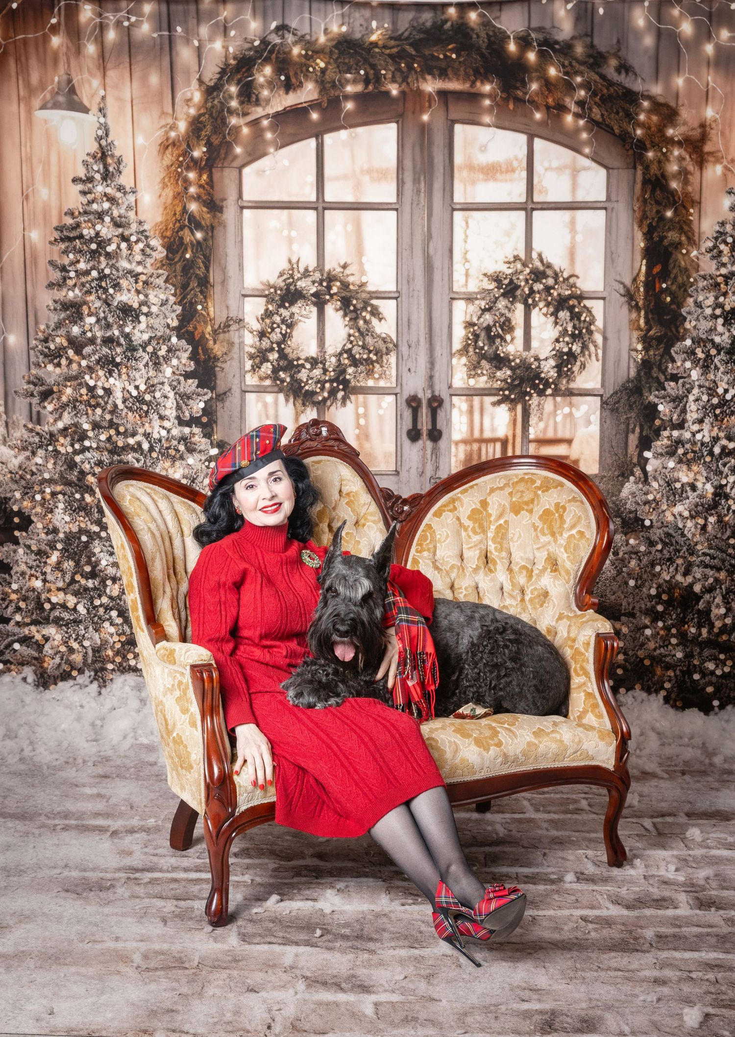 Woman in red dress and hat poses with black Scottish Terrier on vintage sofa amid Christmas trees and wreaths.