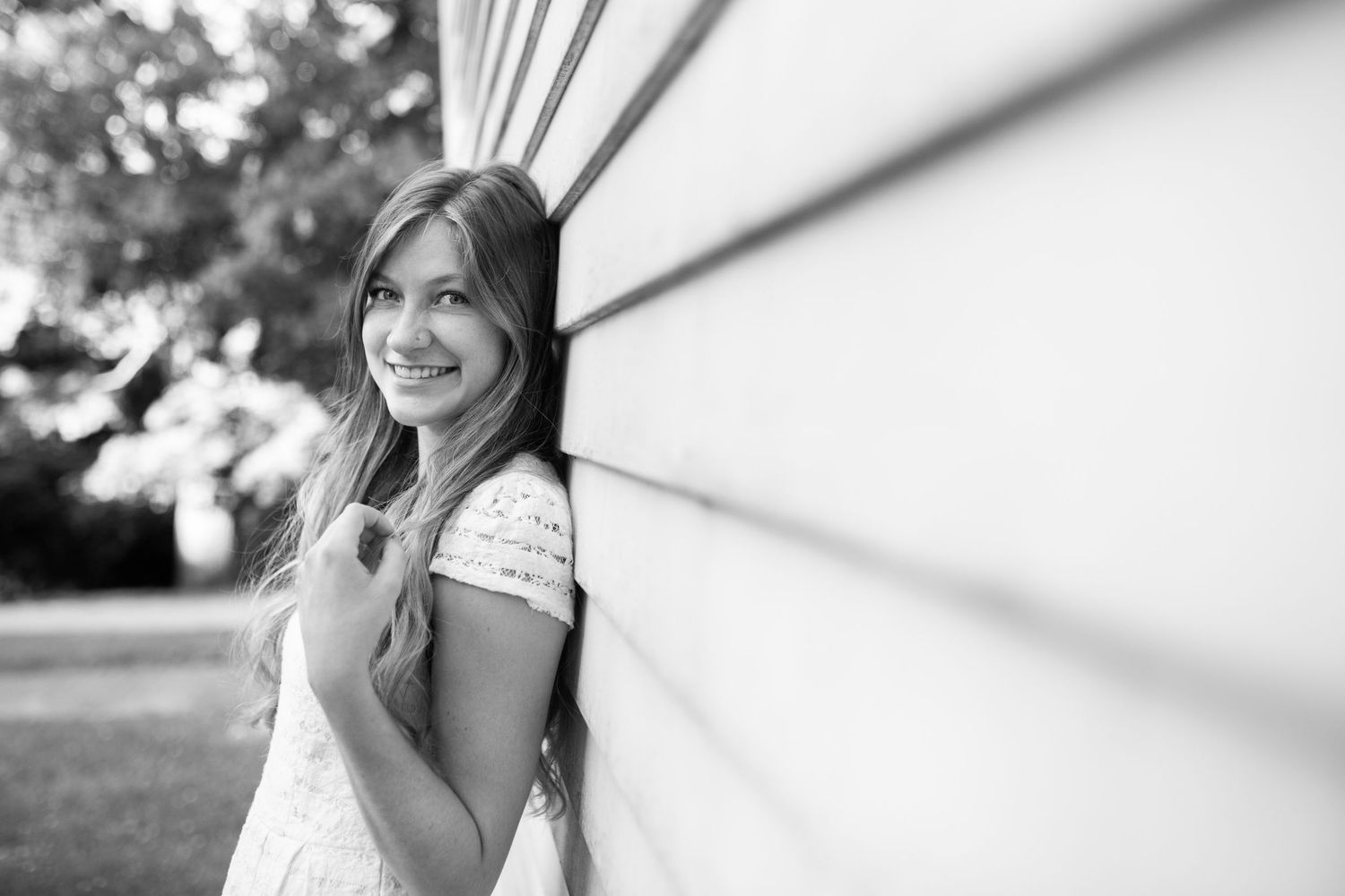 Young woman with long hair smiling and leaning against a white wall outdoors in a black and white portrait photo.