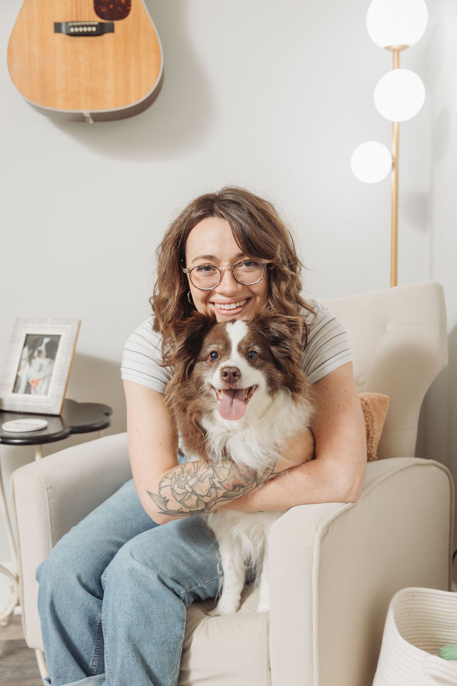 A small brown and white dog sits on a beige couch next to someone in casual attire and glasses.