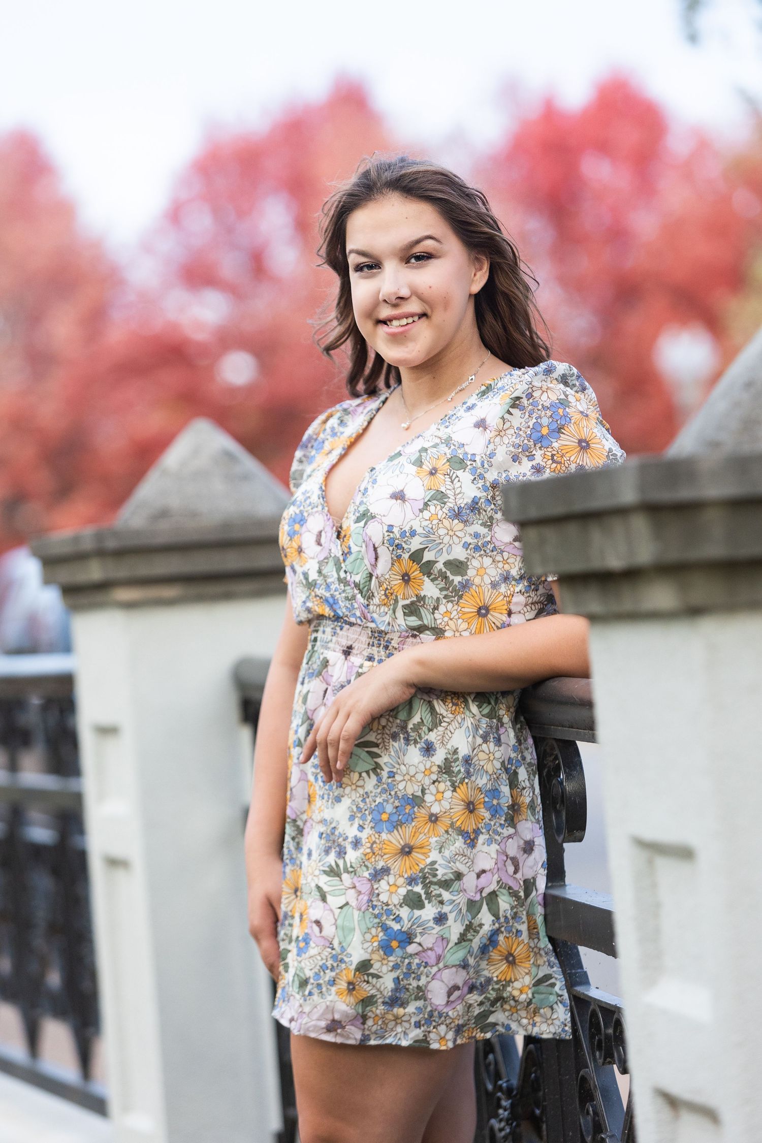 Young woman in floral dress smiling outdoors near a railing with vibrant red autumn trees in the background.